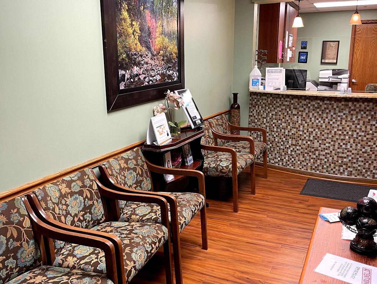 Waiting room with floral cushioned chairs, wooden floor, reception desk, and nature painting on green wall.
